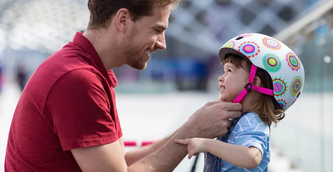 Father smiling as he fastens a colorful Micro Kickboard helmet on his young daughter, highlighting the importance of safety and protective gear.