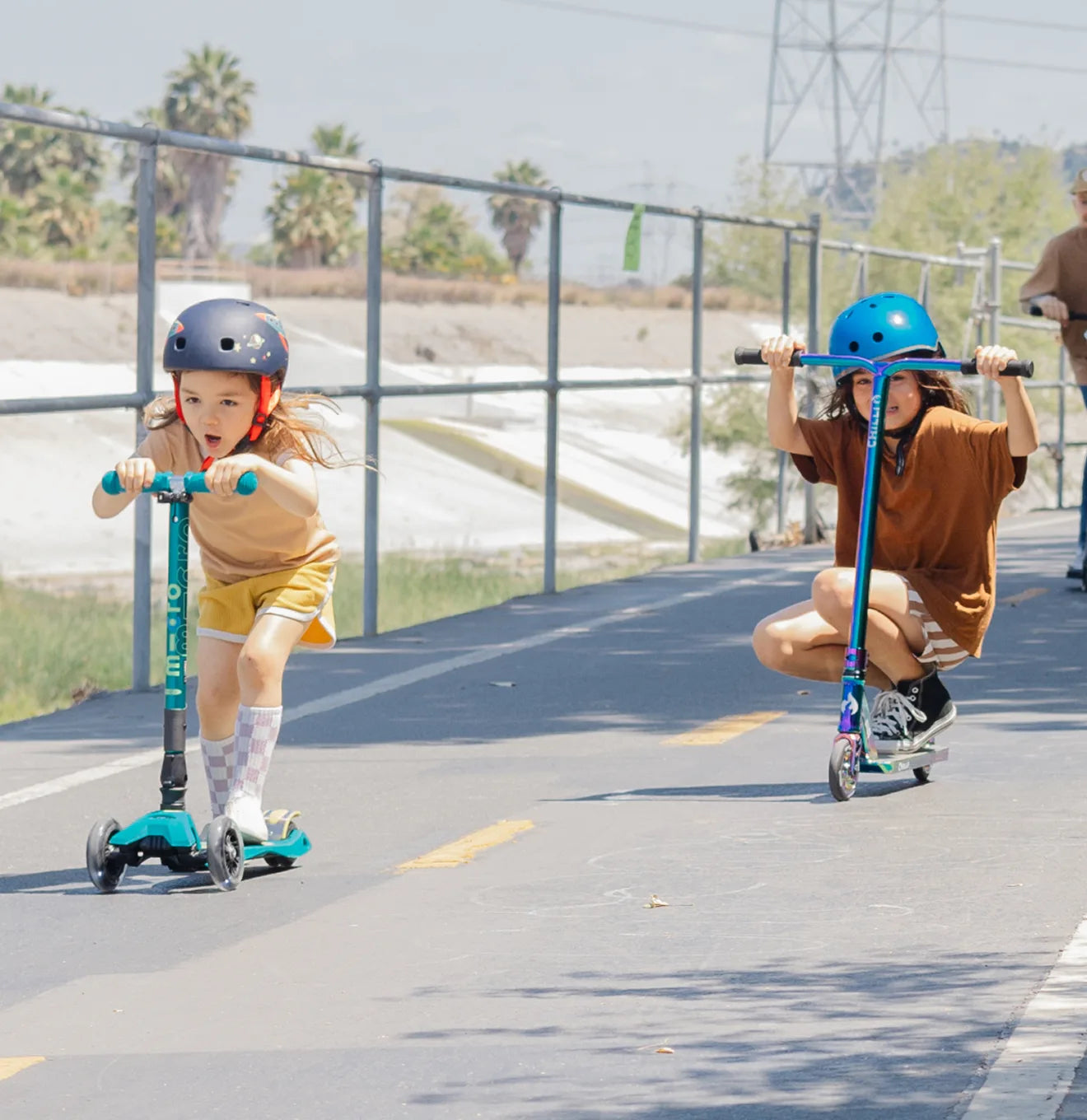 Two young girls riding scooters on a paved path—one on a foldable Micro Kickboard LED 3-wheeled scooter and the other on a Chilli Pro Scooters neochrome 2-wheeled scooter—both wearing helmets for safety and enjoying an energetic outdoor ride.