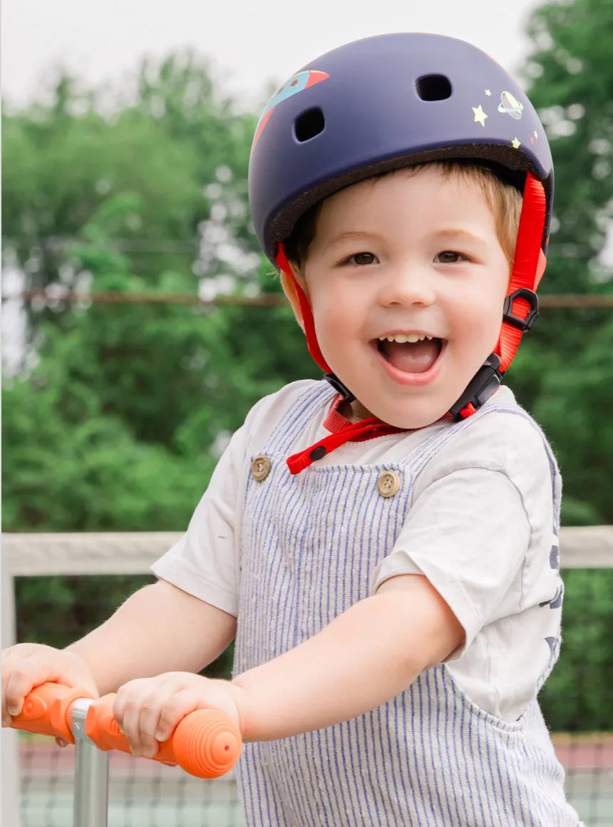 Young child riding a Micro Kickboard Mini 3-wheeled scooter on a sidewalk, wearing a helmet — designed for toddlers and little kids.