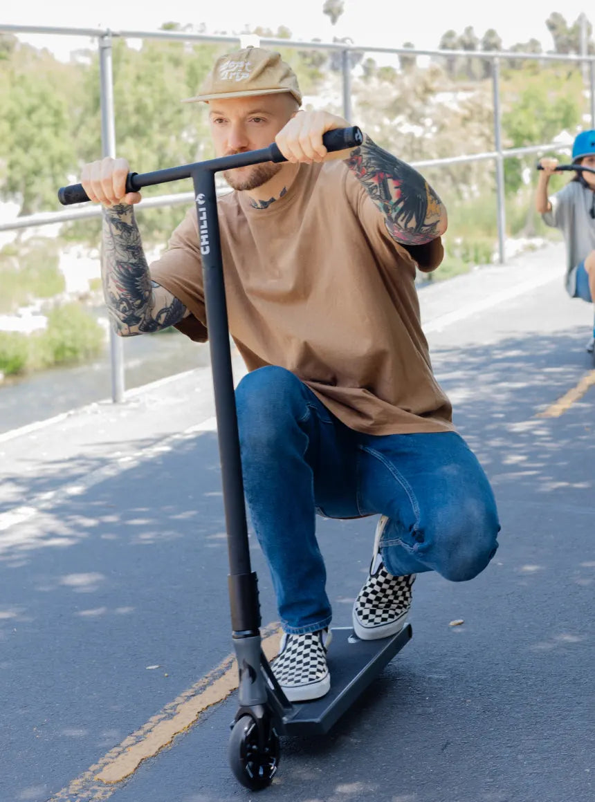 Father rides a black Chilli Zeppelin trick scooter on a paved path, with his young daughter trailing behind; ideal for teens and adults seeking advanced performance.
