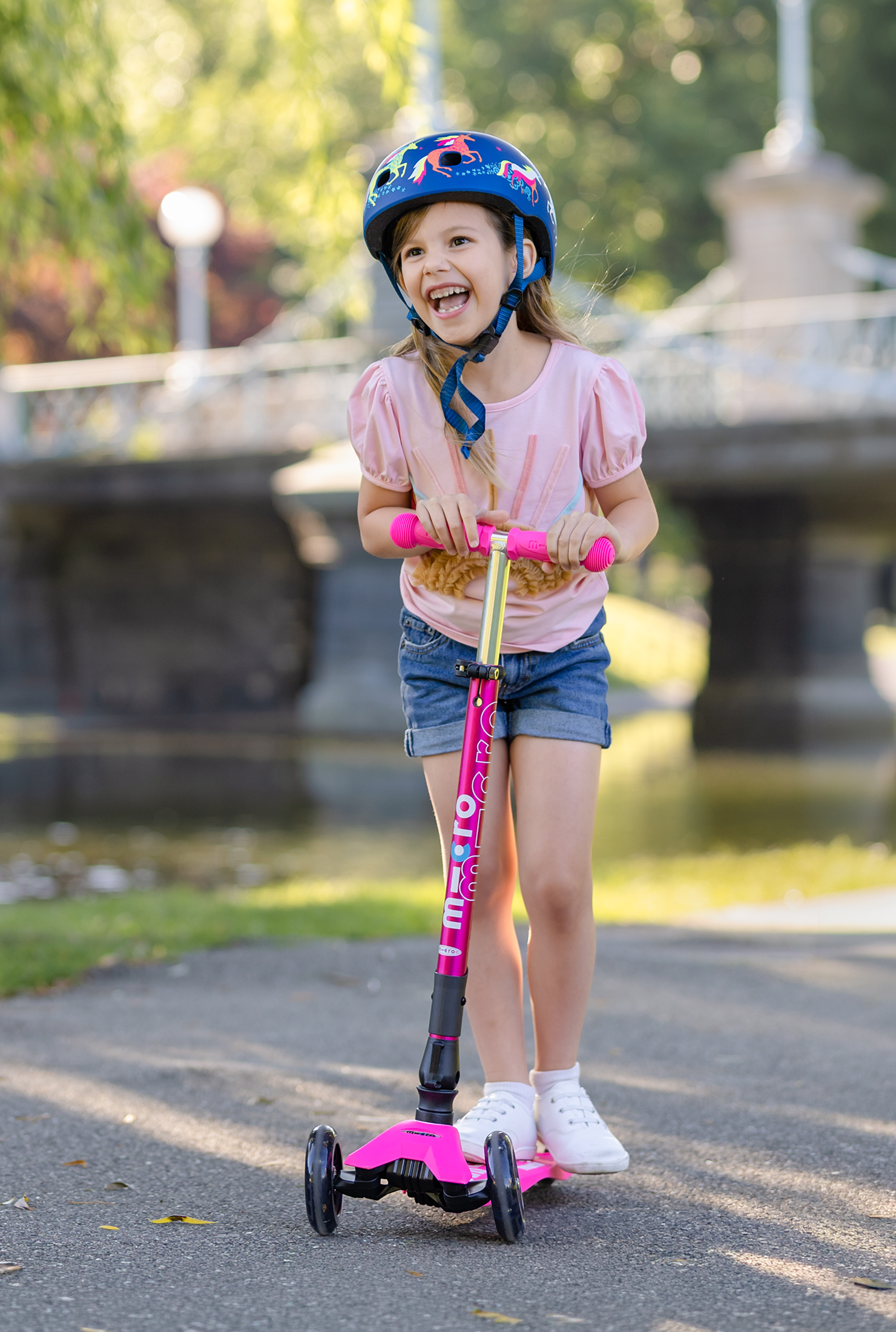 A young girl wearing a blue helmet and a pink t-shirt rides a pink and black three-wheel scooter on an outdoor paved path near a river and bridge.