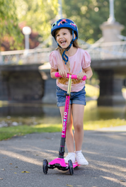 A young girl wearing a blue helmet and a pink t-shirt rides a pink and black three-wheel scooter on an outdoor paved path near a river and bridge.